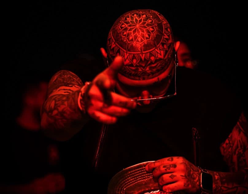A tattooed man lit in deep red, hands raised to his head, with an intricate mandala tattoo across his shaved scalp.