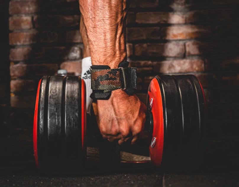 A veined forearm in a lifting strap gripping a loaded dumbbell against a brick wall.