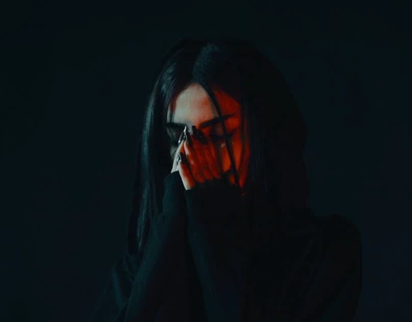 A hooded figure in dark sunglasses photographed against black, with hair falling across the face.