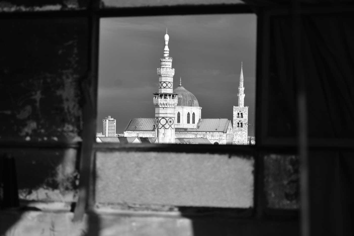 The dome and minarets of the Umayyad Mosque in Damascus, seen in black and white through a derelict window.